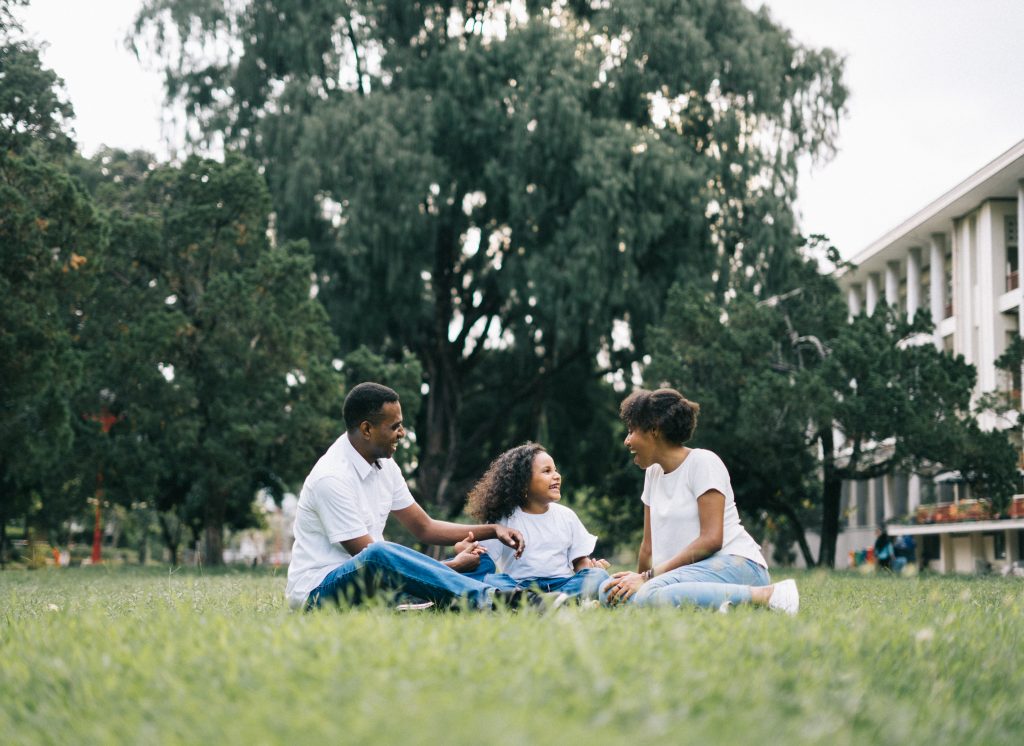 Two parents and their child sitting in a grassy park and laughing.