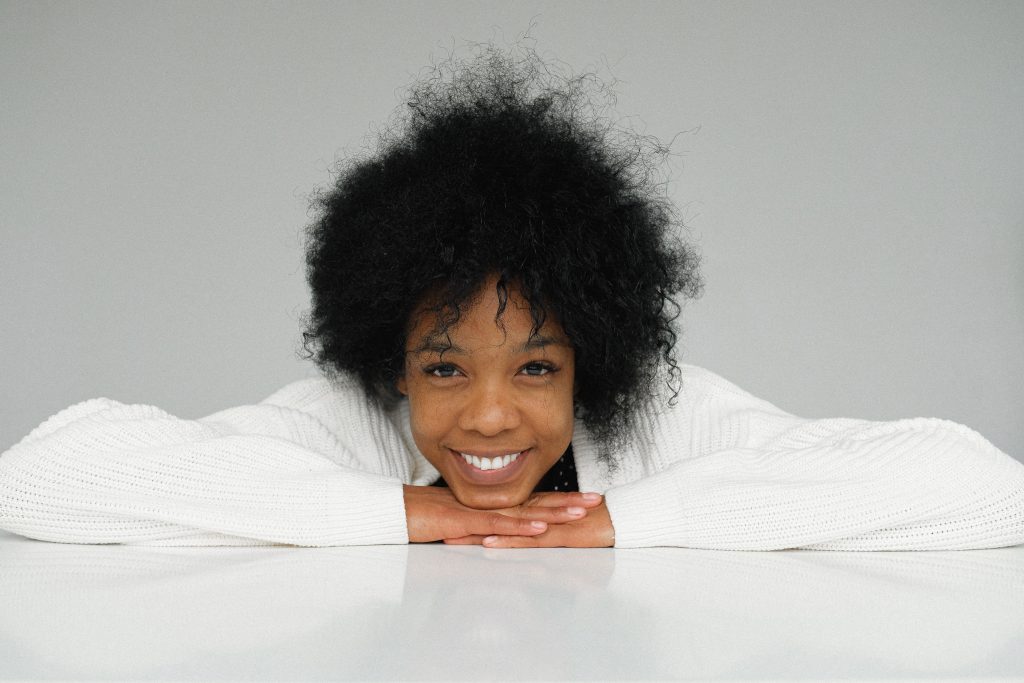 Smiling young woman resting her chin atop her hands, which are lying flat on a table.